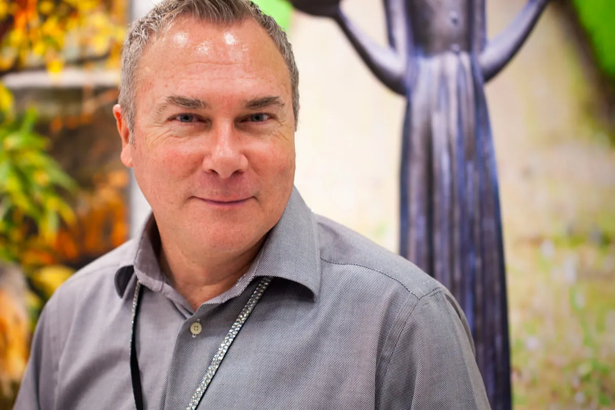 Portrait of Brian in a grey shirt outdoors, a carved wooden figure blurred behind