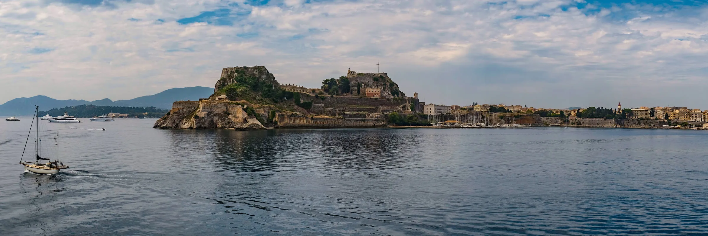 The Old Fortress of Corfu on its headland under bright cloud, a sailing boat crossing in front