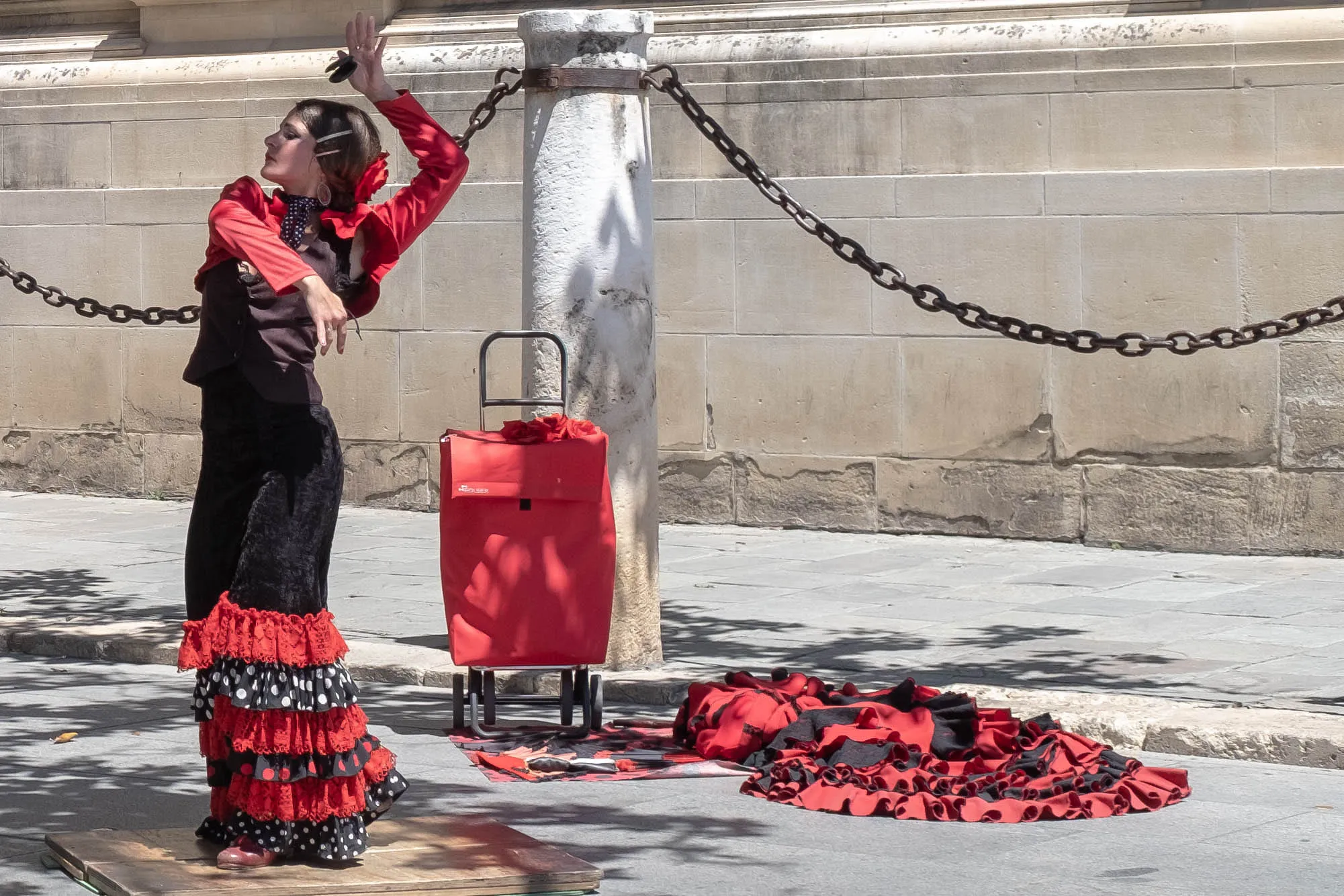 A flamenco dancer mid-turn on a wooden board in the street, arms raised, red fabric at her feet