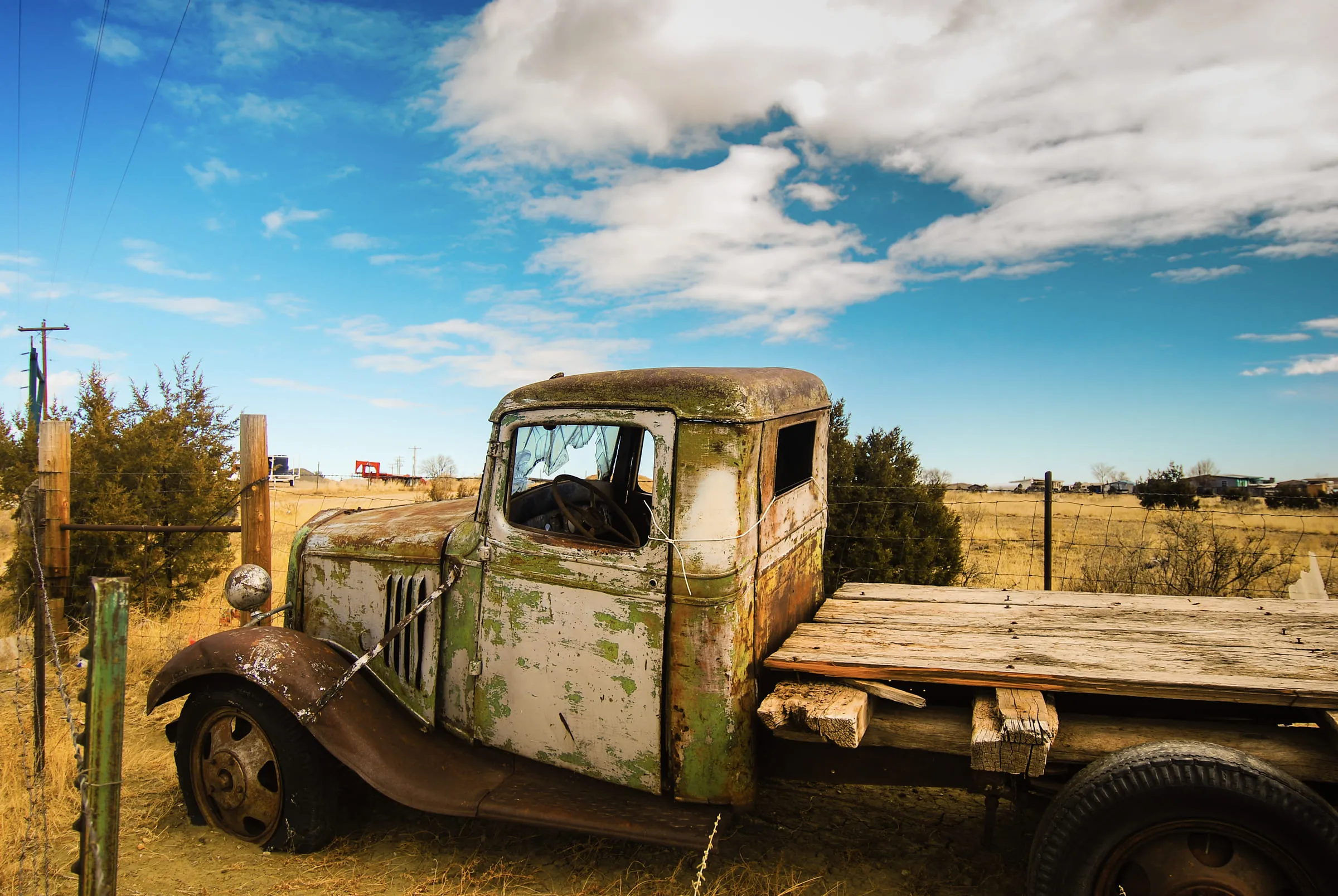 A rusted 1930s flatbed truck with peeling green paint, abandoned in a field under a bright sky
