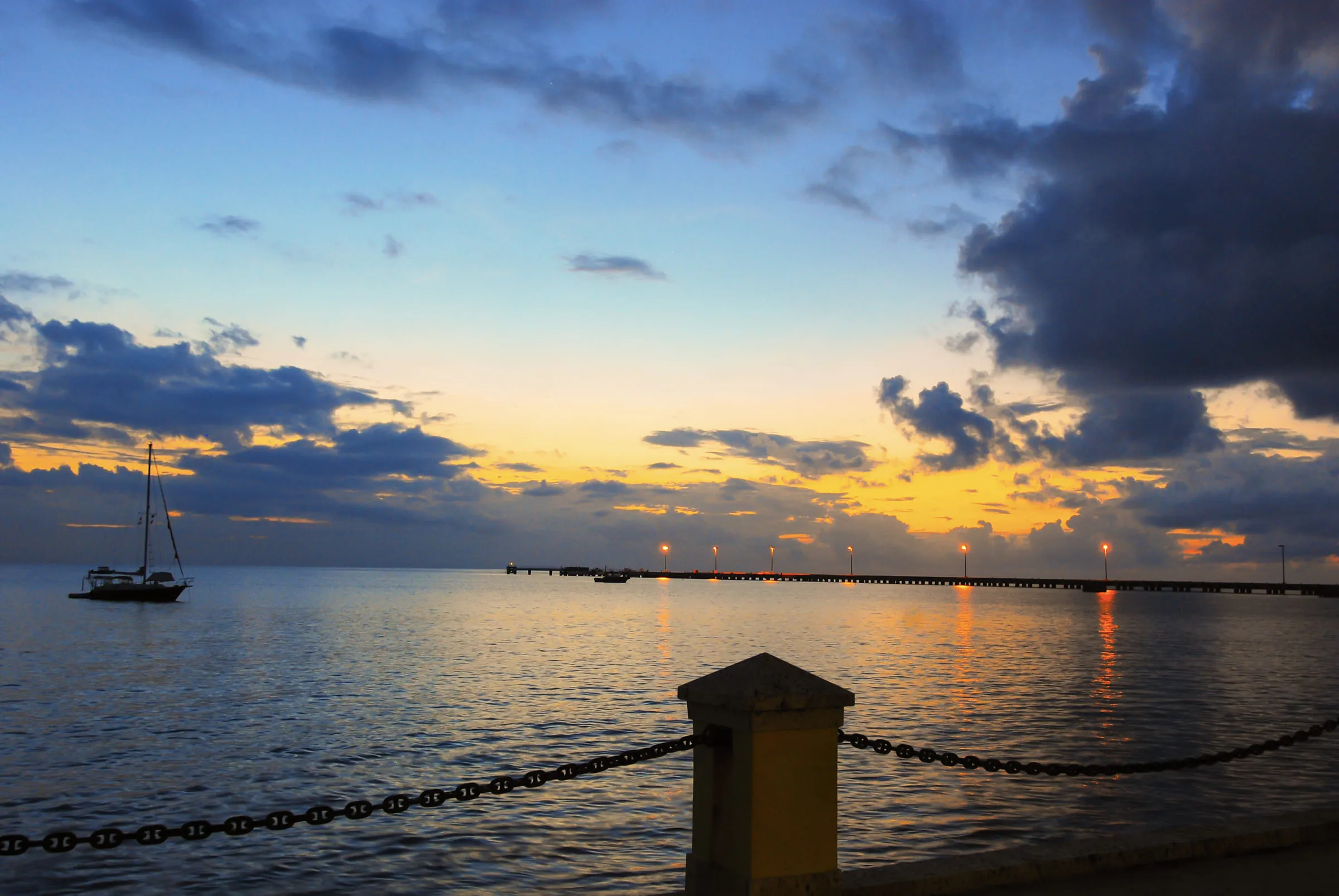 A sailing boat moored off a pier at dusk, harbour lights reflected in still water