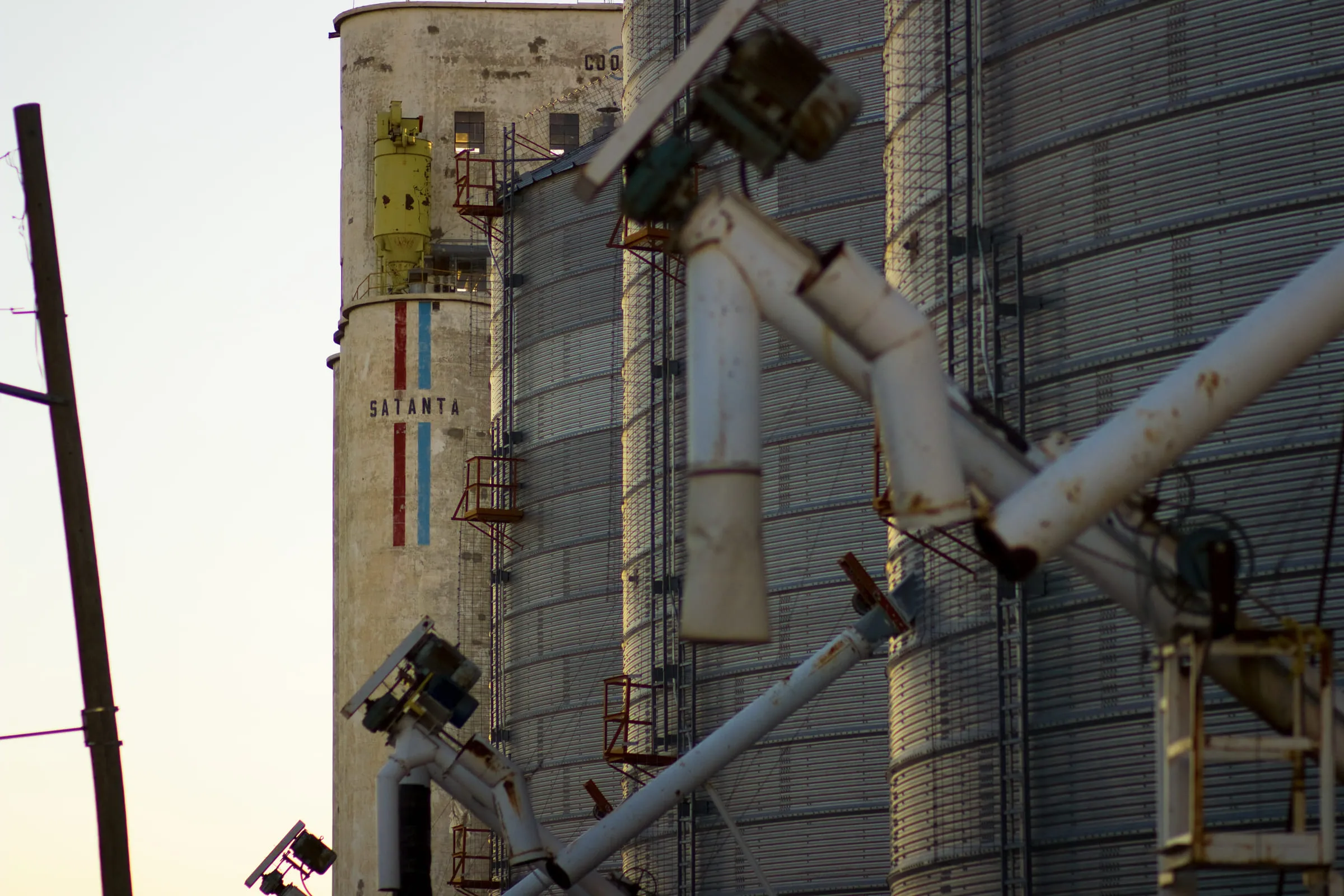 Steel grain bins and conveyor pipes lit warm at low sun, the word SATANTA on the tower