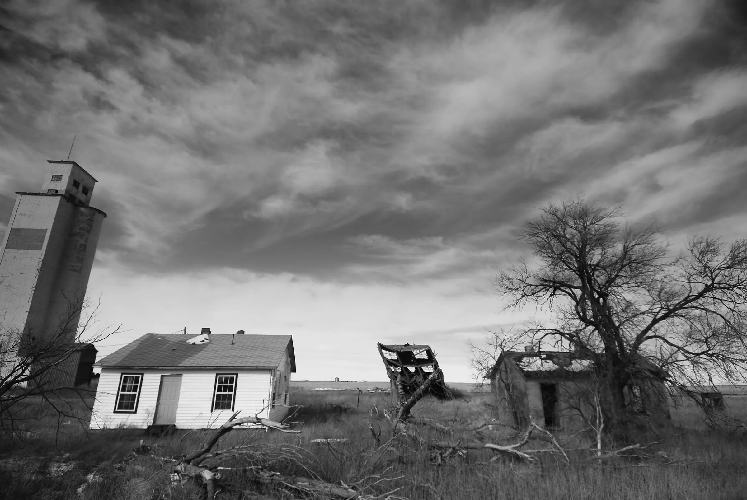 Black and white view of an abandoned farmhouse and outbuildings under a dramatic streaked sky