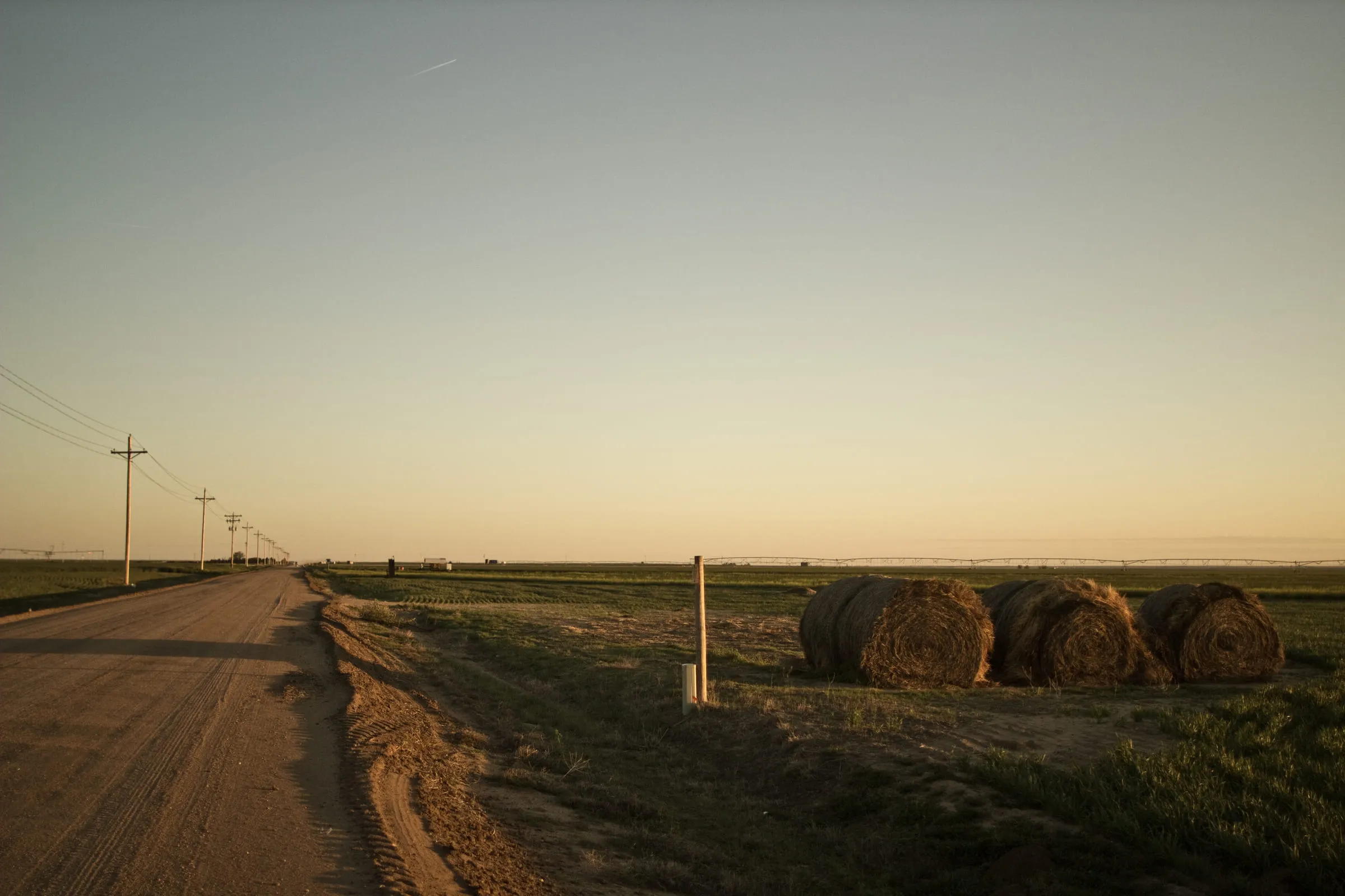 A dirt road running beside a fence line and round hay bales at dusk, power poles receding