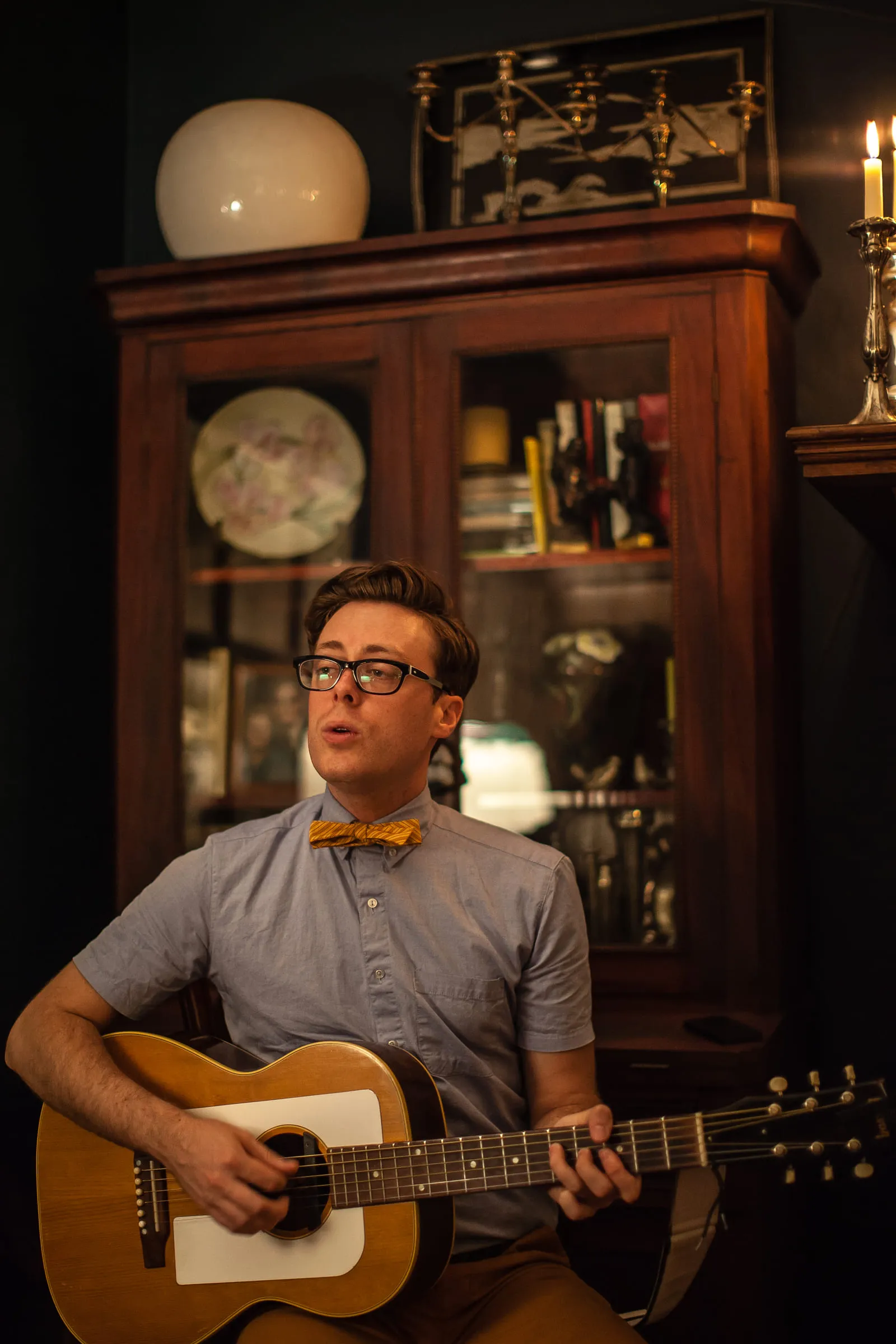 Jeremy Messersmith seated with an acoustic guitar in a room with a tall wooden cabinet