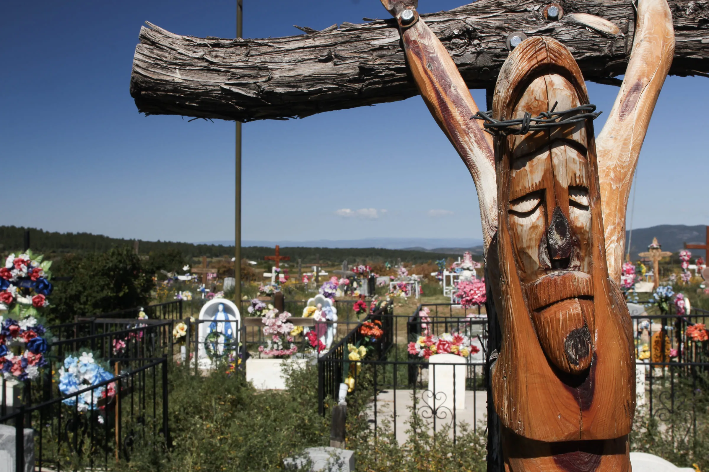 A carved wooden crucifix in the foreground of a cemetery filled with bright artificial flowers