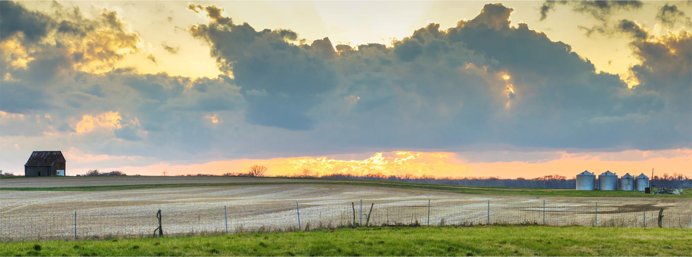 A barn and grain bins across a flat field at sunset, a long low cloud bank catching the light
