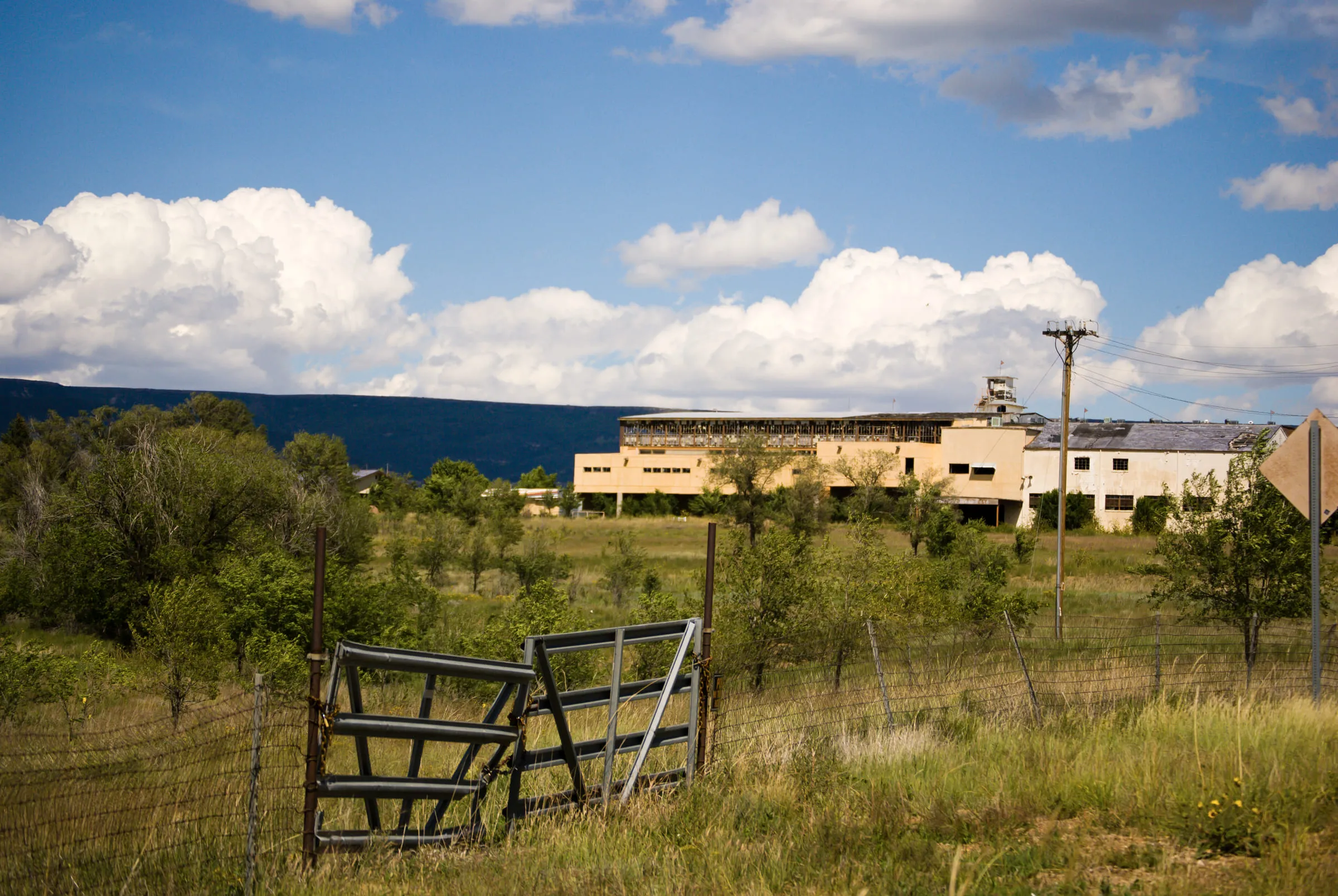 A broken metal gate hanging open in a fence, a long derelict grandstand building beyond it