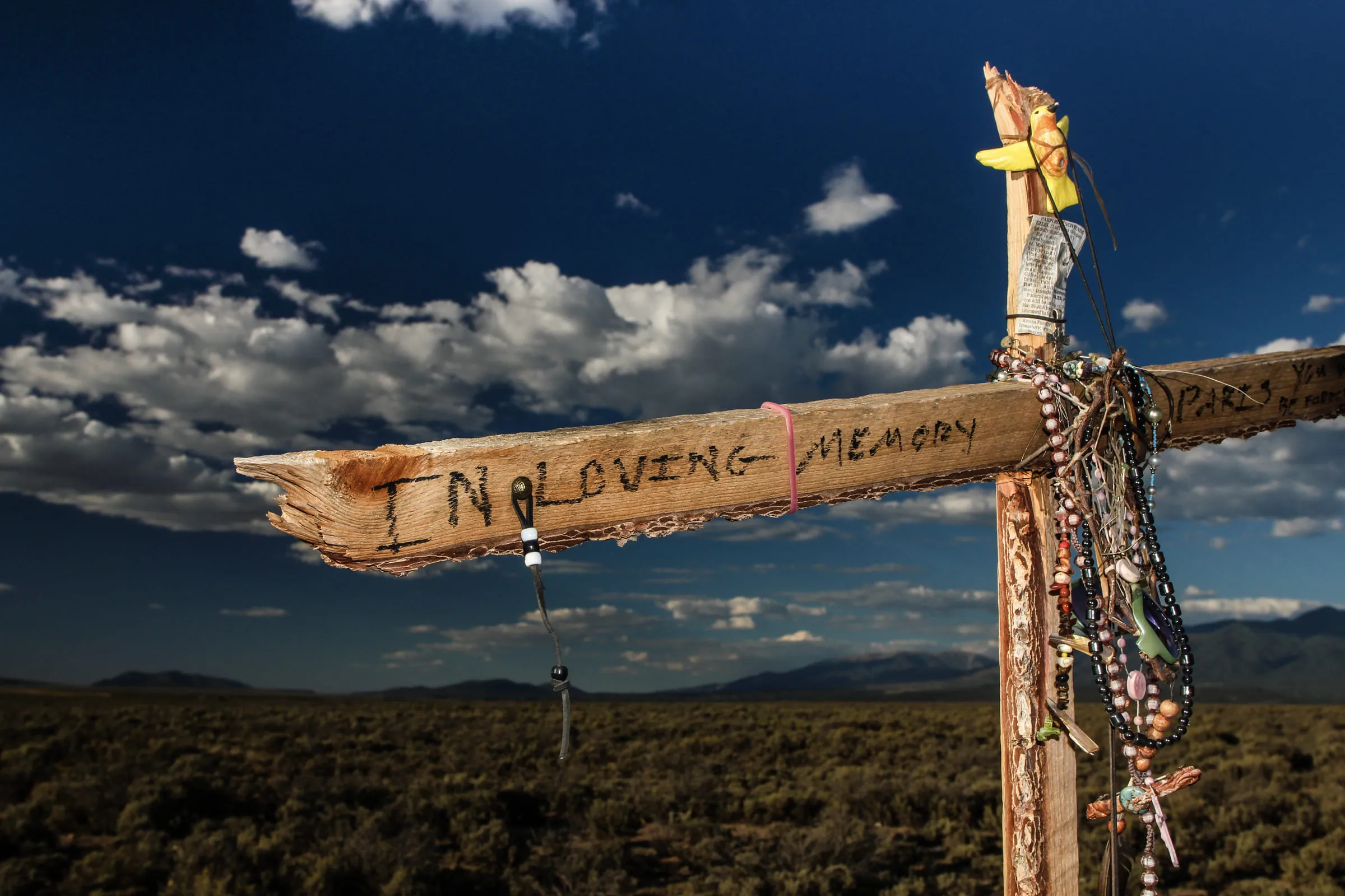 A roadside memorial cross reading "In Loving Memory", hung with beads and ribbon, against evening desert