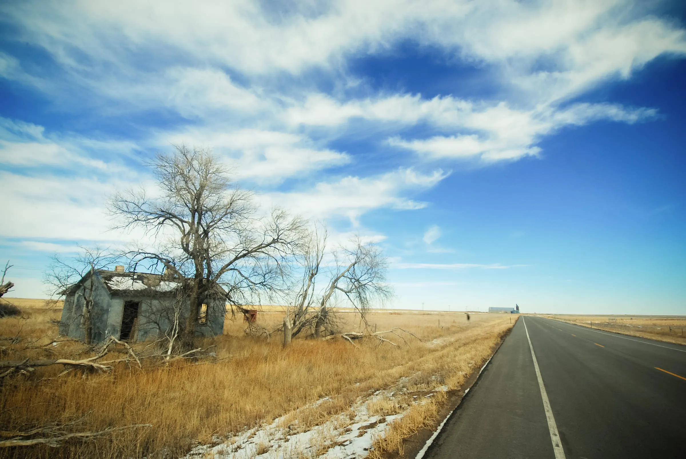 An empty two-lane highway running past a derelict stone house and bare trees under a wide blue sky