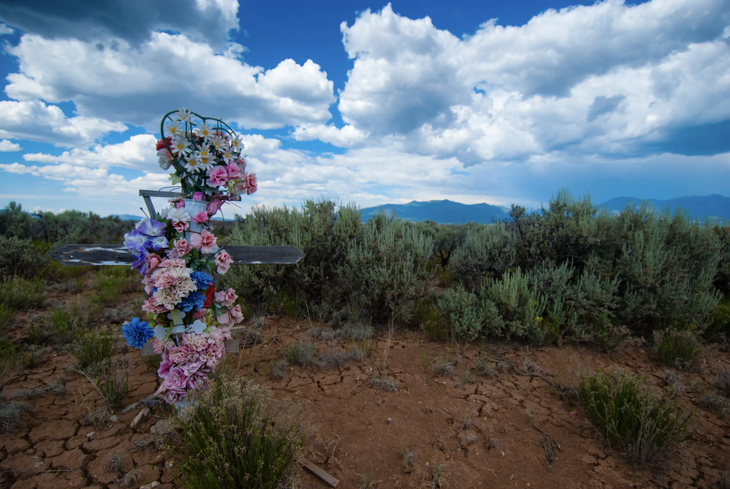 A roadside cross wrapped in pink and blue artificial flowers, standing in sagebrush under big clouds