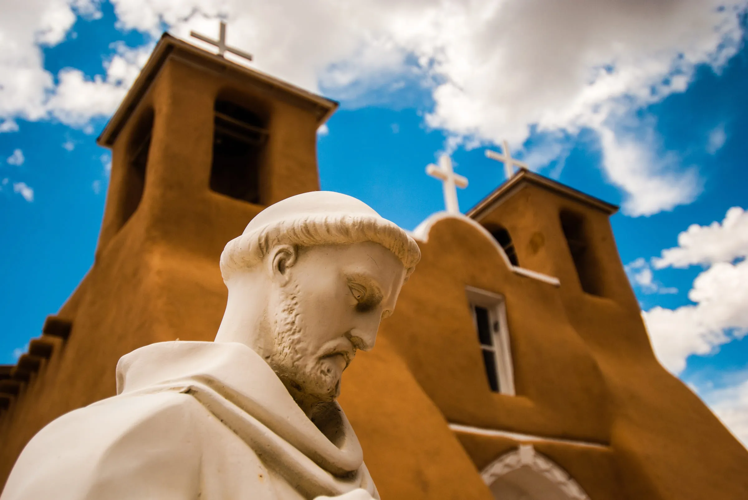 A white stone statue of Saint Francis in front of the twin adobe bell towers of San Francisco de Asís