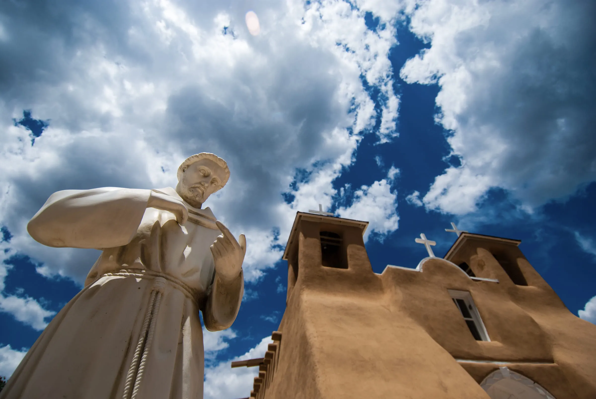 The Saint Francis statue and adobe church seen from below against towering white cloud and blue sky