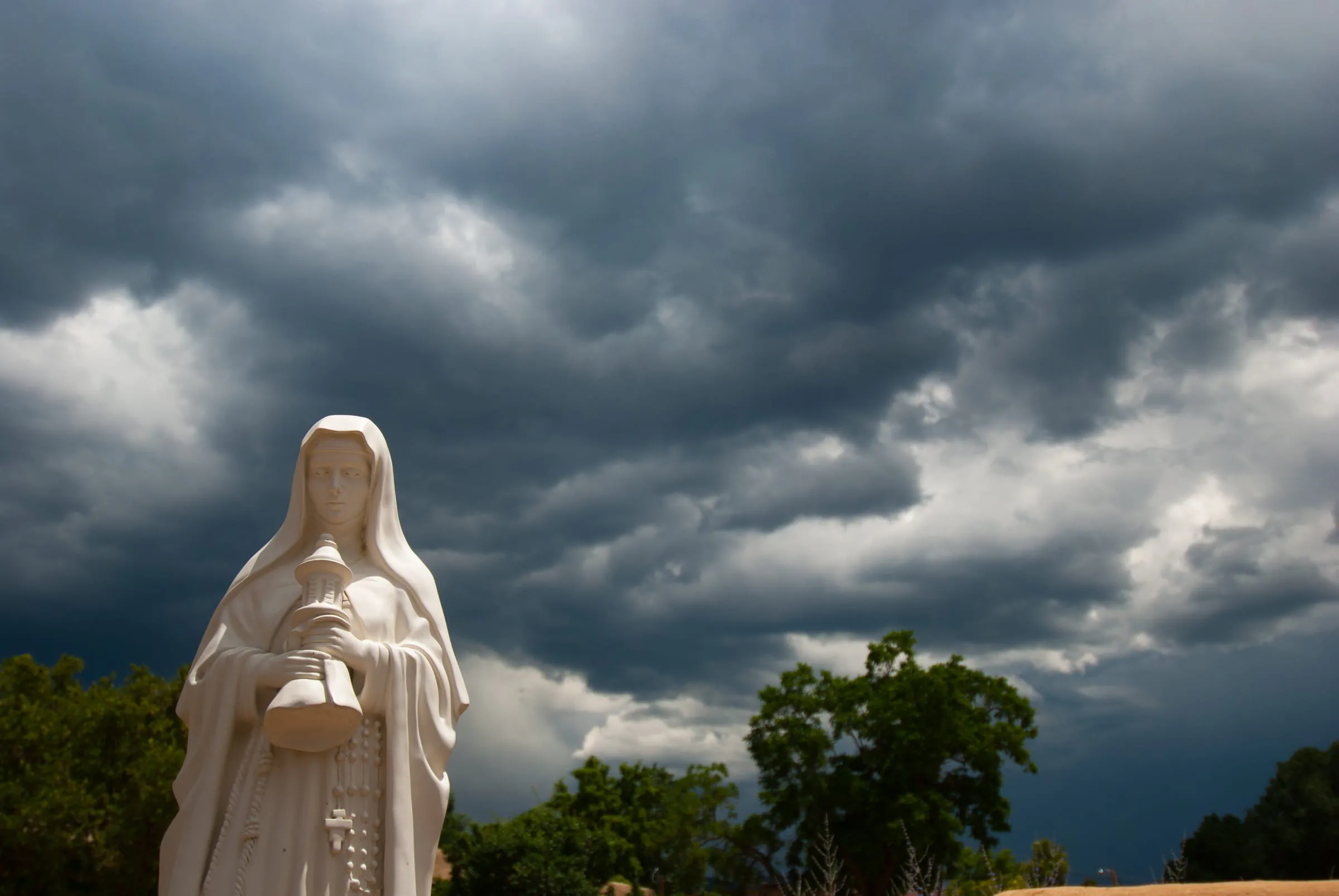 A white statue of a veiled woman holding a basket, silhouetted against a bank of storm cloud
