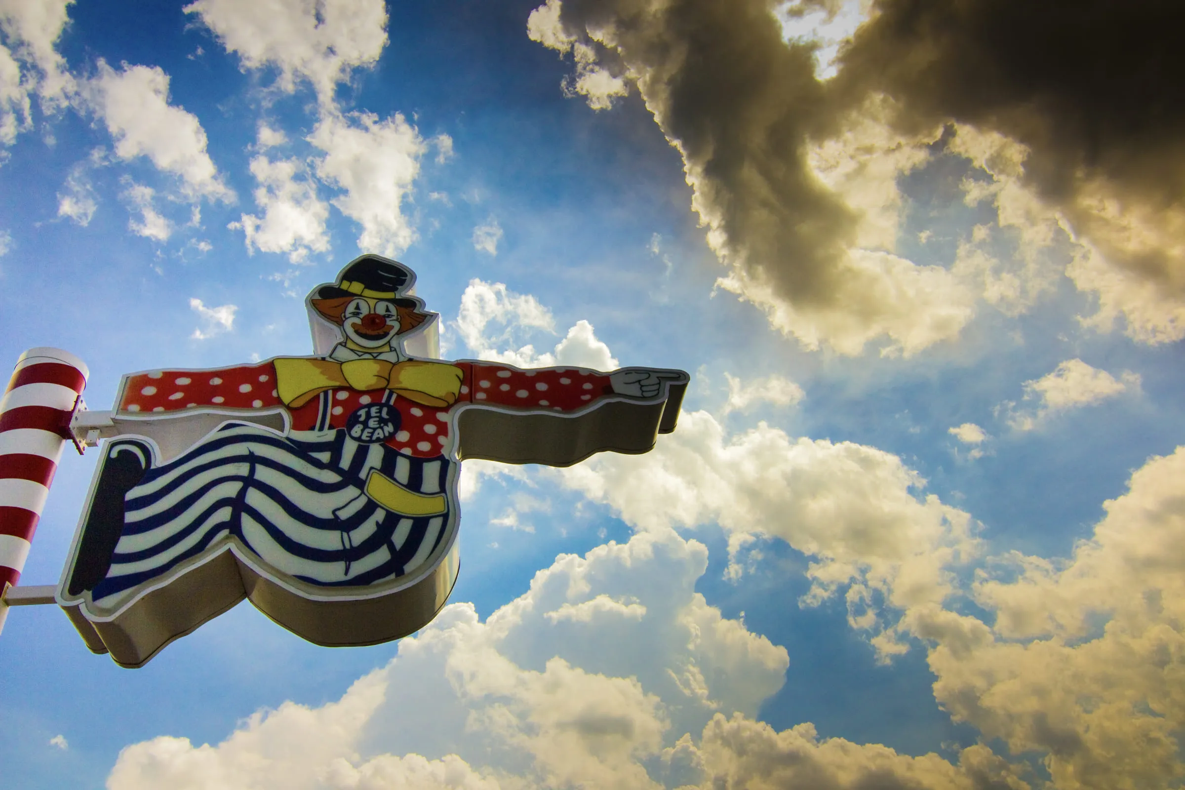 A painted metal clown sign shot from directly below against piled clouds and blue sky