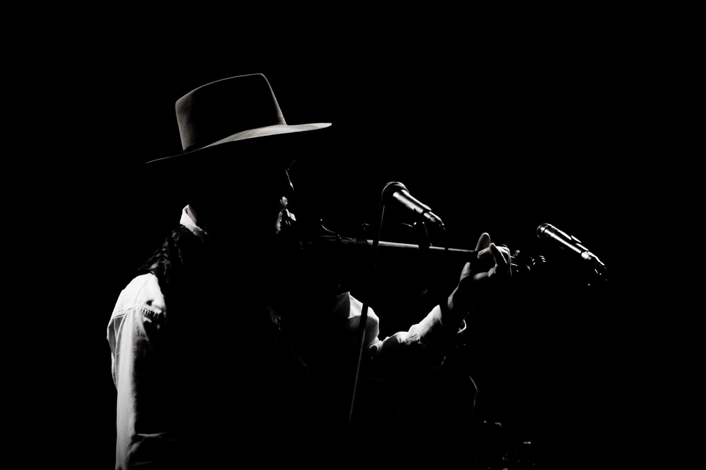 A musician in a wide-brimmed hat playing at a microphone, lit hard against a black background