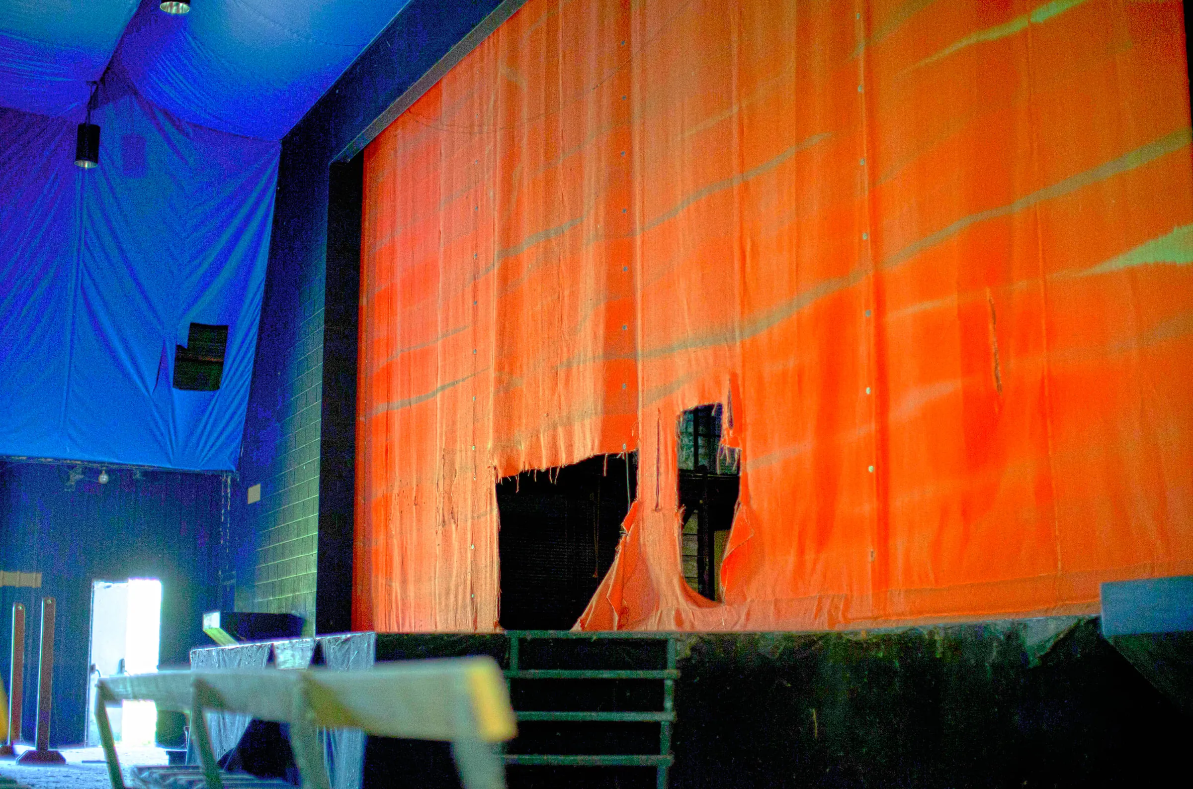 The same torn orange curtain in brighter light, stage steps and scattered furniture below it