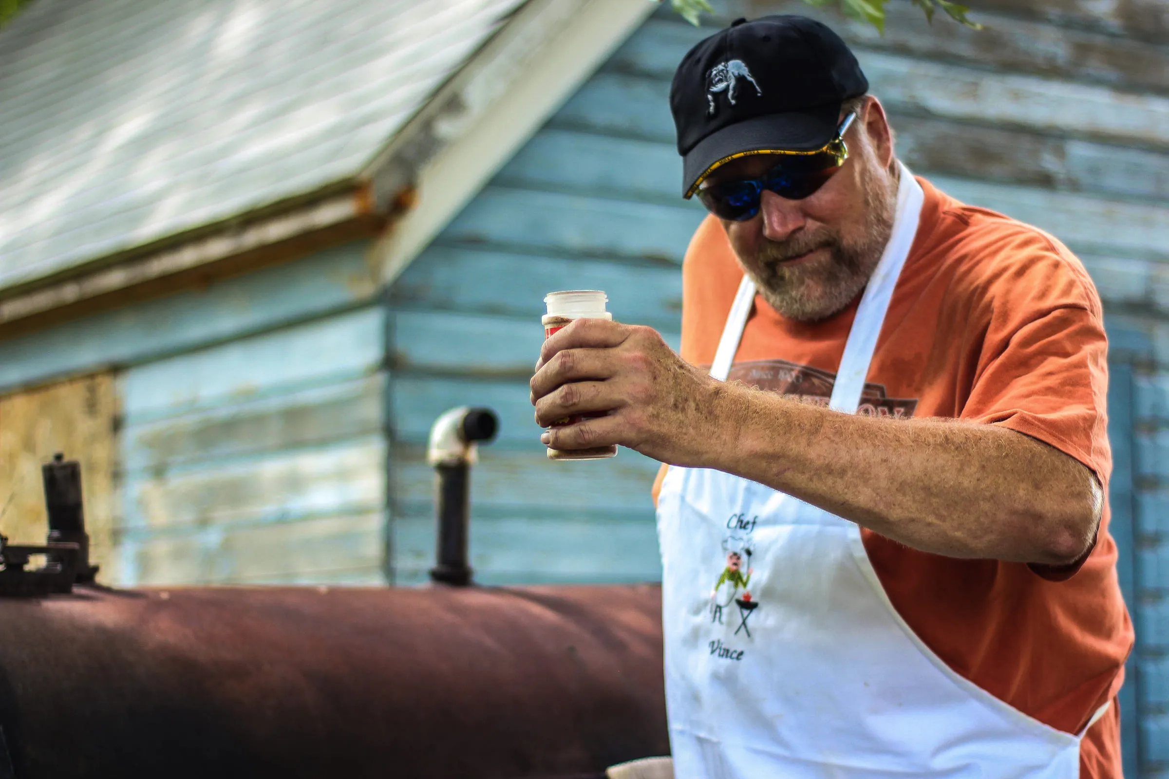 Vince in an apron and cap holding a drink beside a barbecue smoker, blue clapboard wall behind